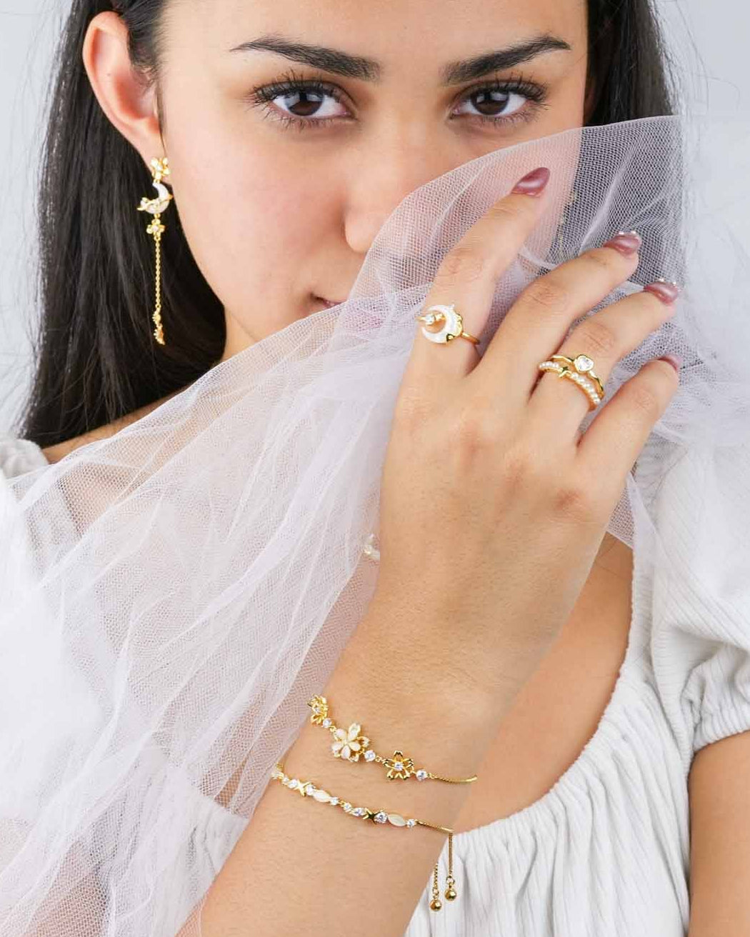 Woman wearing gold jewelry including earrings, rings, and bracelets, holding a white scarf.