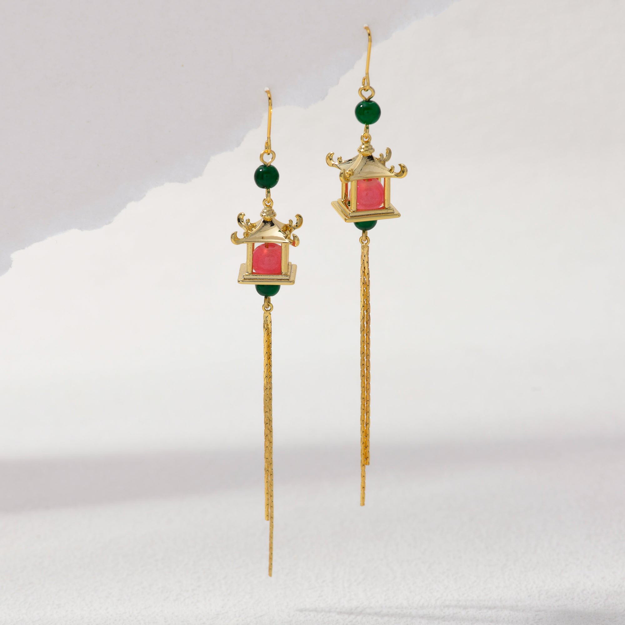 A pair of gold dangle earrings with green bead accents, a small lantern-shaped pink and green centerpiece, and long dangling chains, shown on a light gray background.