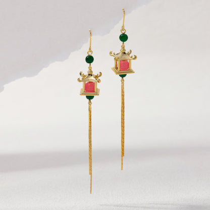 A pair of gold dangle earrings with green bead accents, a small lantern-shaped pink and green centerpiece, and long dangling chains, shown on a light gray background.